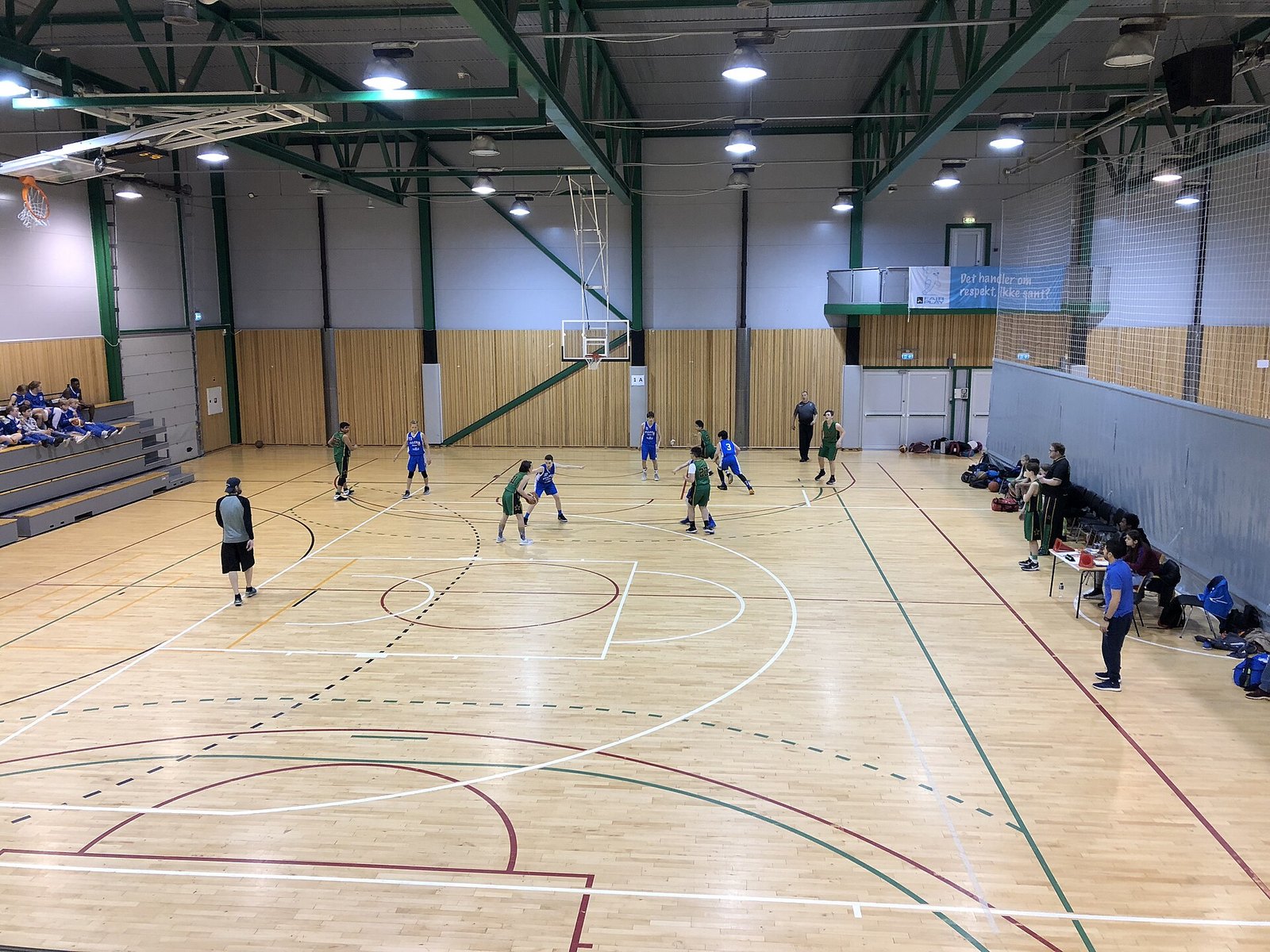 Inomhushall med basketmatch på parkettgolv sedd från läktaren, med korgar och strålkastare i taket